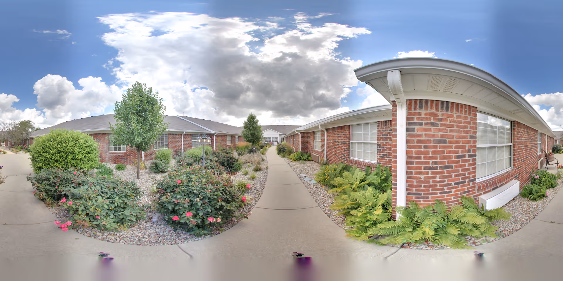 Outdoor walkway between two single-story brick buildings with landscaped garden beds containing bushes, flowers, and small trees under a partly cloudy sky.