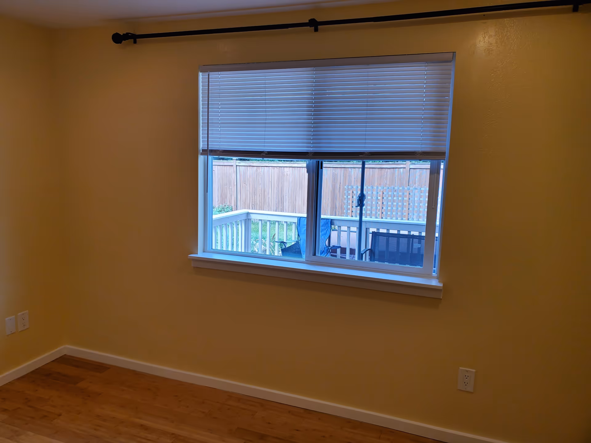 Empty room with yellow walls, wooden floor, a window with partially closed blinds showing a fenced backyard with patio chairs outside.