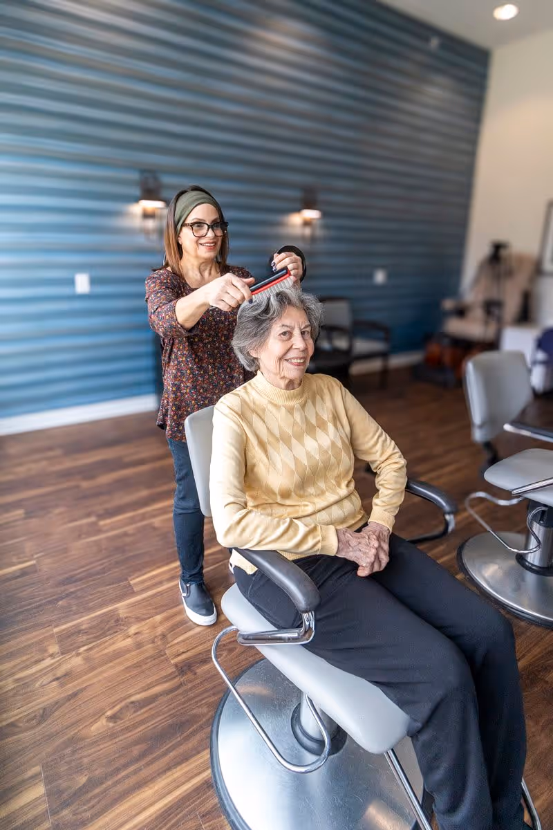 An elderly woman sitting in a salon chair smiling while a hairstylist combs her gray hair in a modern salon with wooden floors and blue paneled walls.