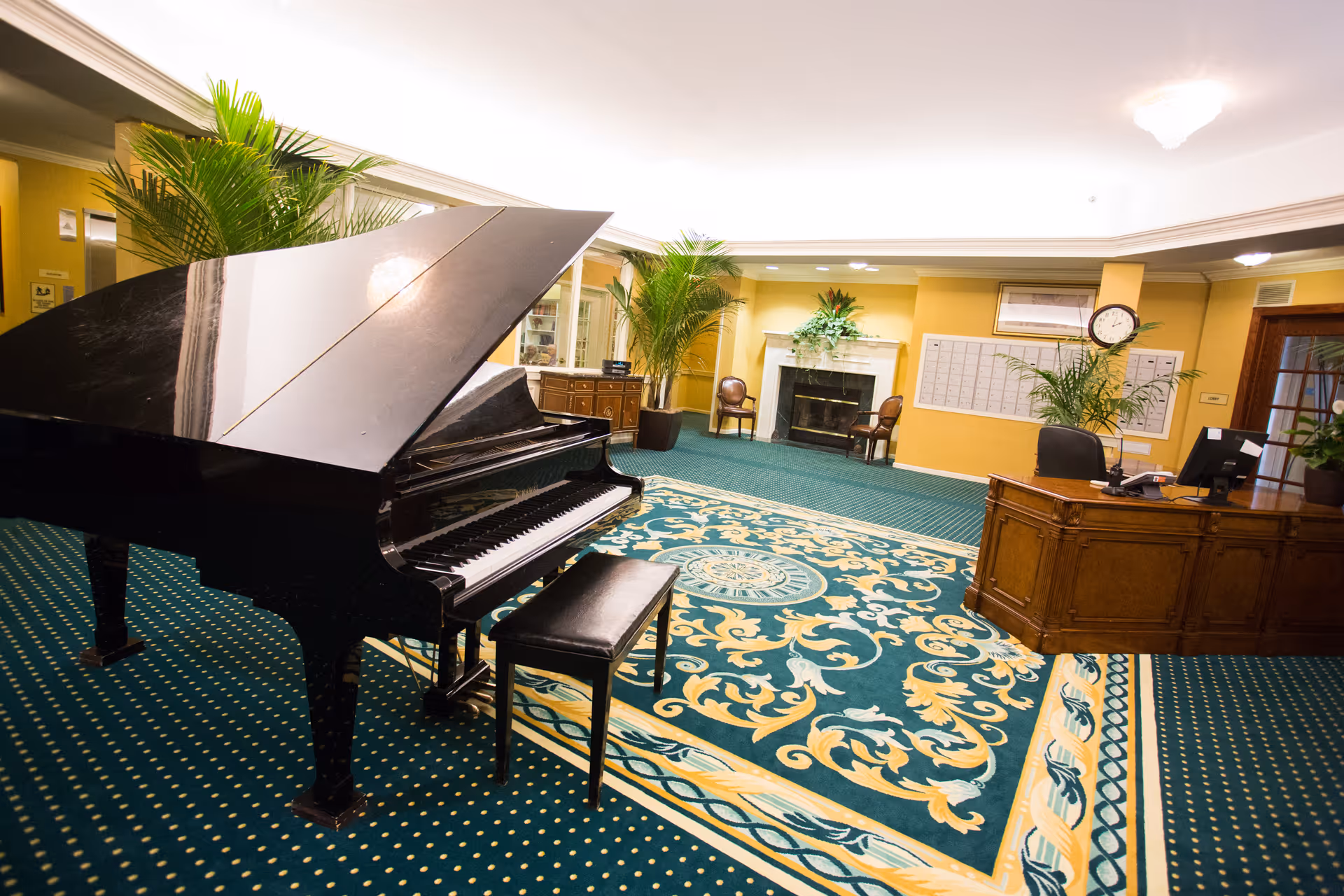 Lobby interior with a black grand piano, patterned carpet, reception desk, fireplace and potted plants.