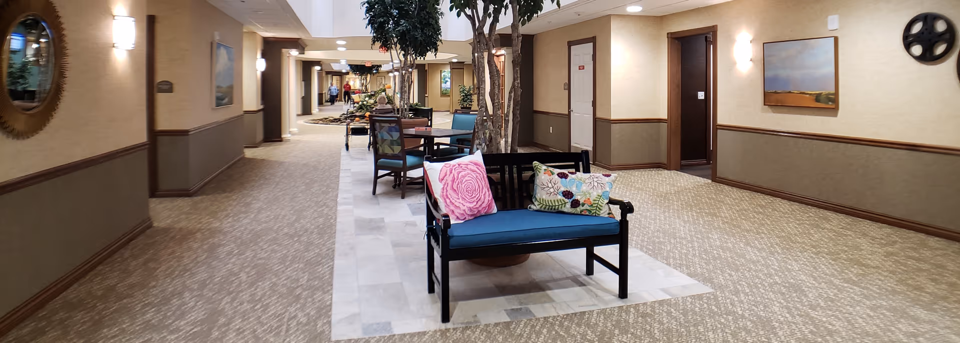 A wide hallway in a retirement community with carpeted floors and beige walls. The hallway features seating areas with chairs and benches adorned with decorative pillows, potted trees, wall-mounted lights, and framed artwork. Doors to rooms line the corridor, and a person is visible in the distance walking down the hall.