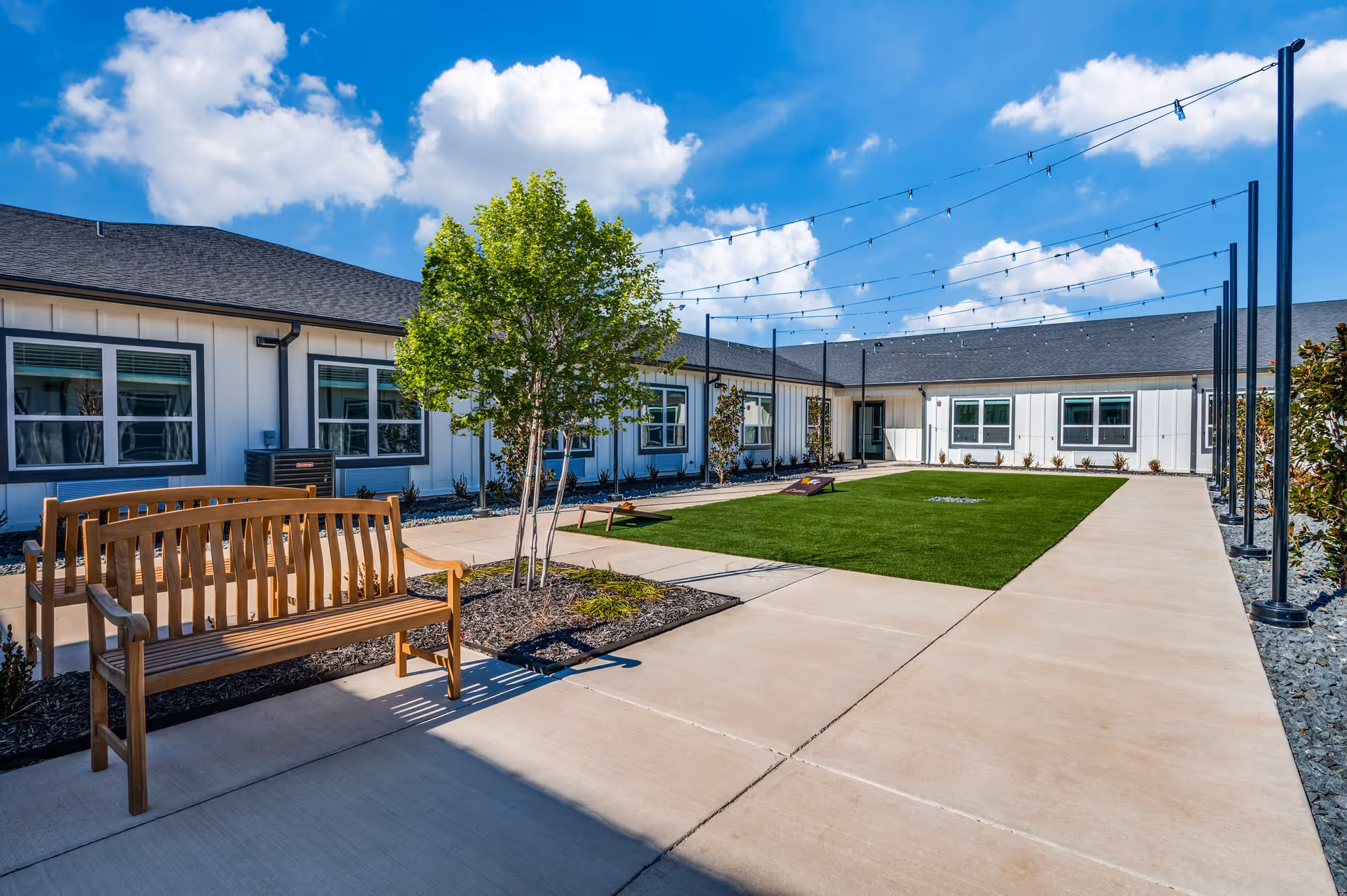 Outdoor courtyard area at The Lodge of Saginaw Health & Wellness featuring a wooden bench, a small tree, string lights overhead, a green lawn with a cornhole game set, and a surrounding building with multiple windows under a bright blue sky with scattered clouds.