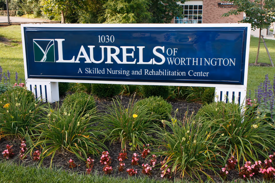 A blue outdoor sign reading "Laurels of Worthington, A Skilled Nursing and Rehabilitation Center" surrounded by landscaped flowers and greenery.