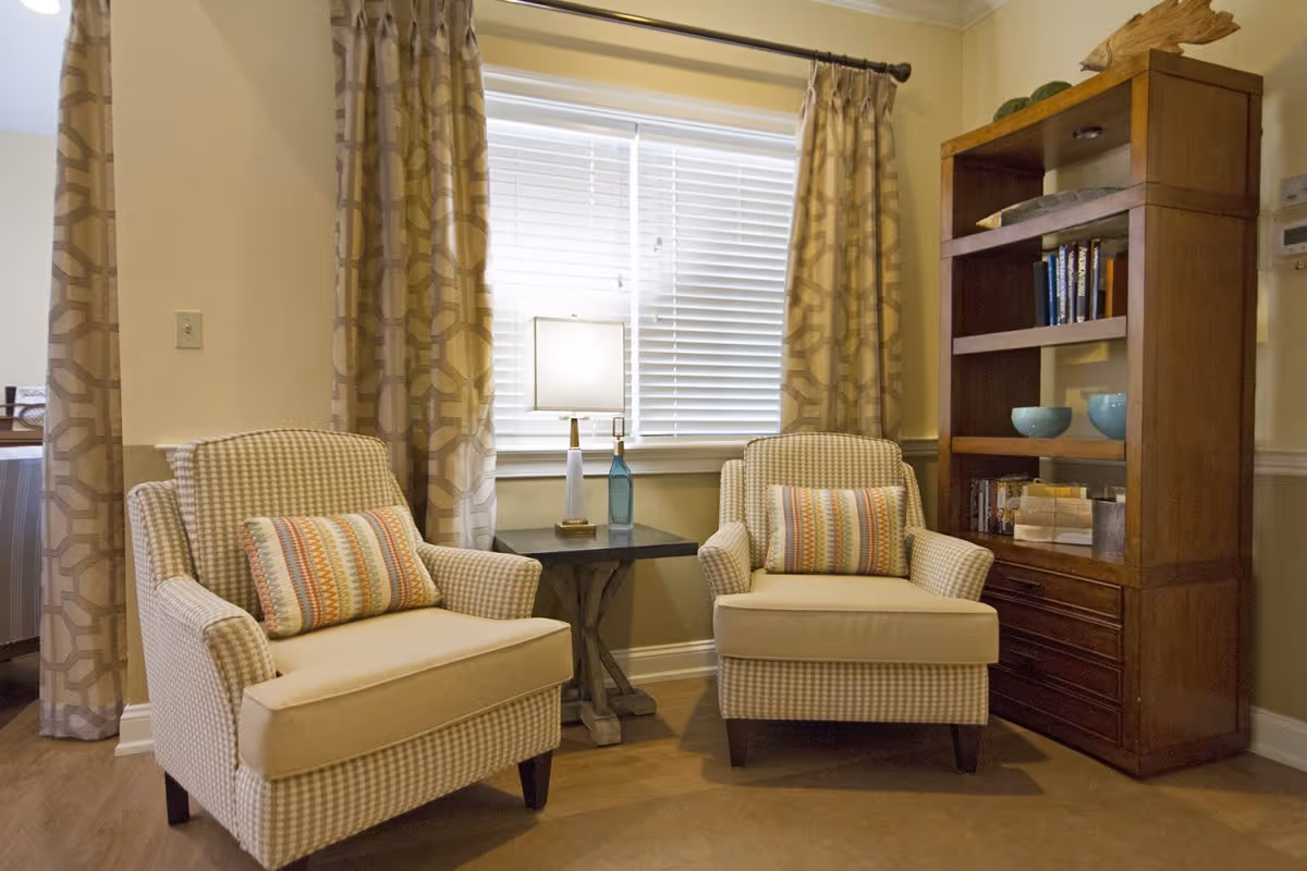 A cozy sitting area with two beige patterned armchairs, each with a colorful striped lumbar pillow. Between the chairs is a small dark wooden side table with a lamp and decorative bottles. Behind the chairs is a window with white blinds and beige patterned curtains. To the right is a tall wooden bookshelf with books and decorative items.