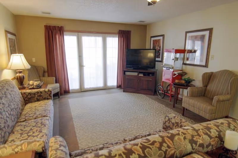 Cozy assisted-living common room with patterned sofas and armchairs arranged around a central rug, a TV on a wooden console, French doors with curtains, and a popcorn machine on a side table.