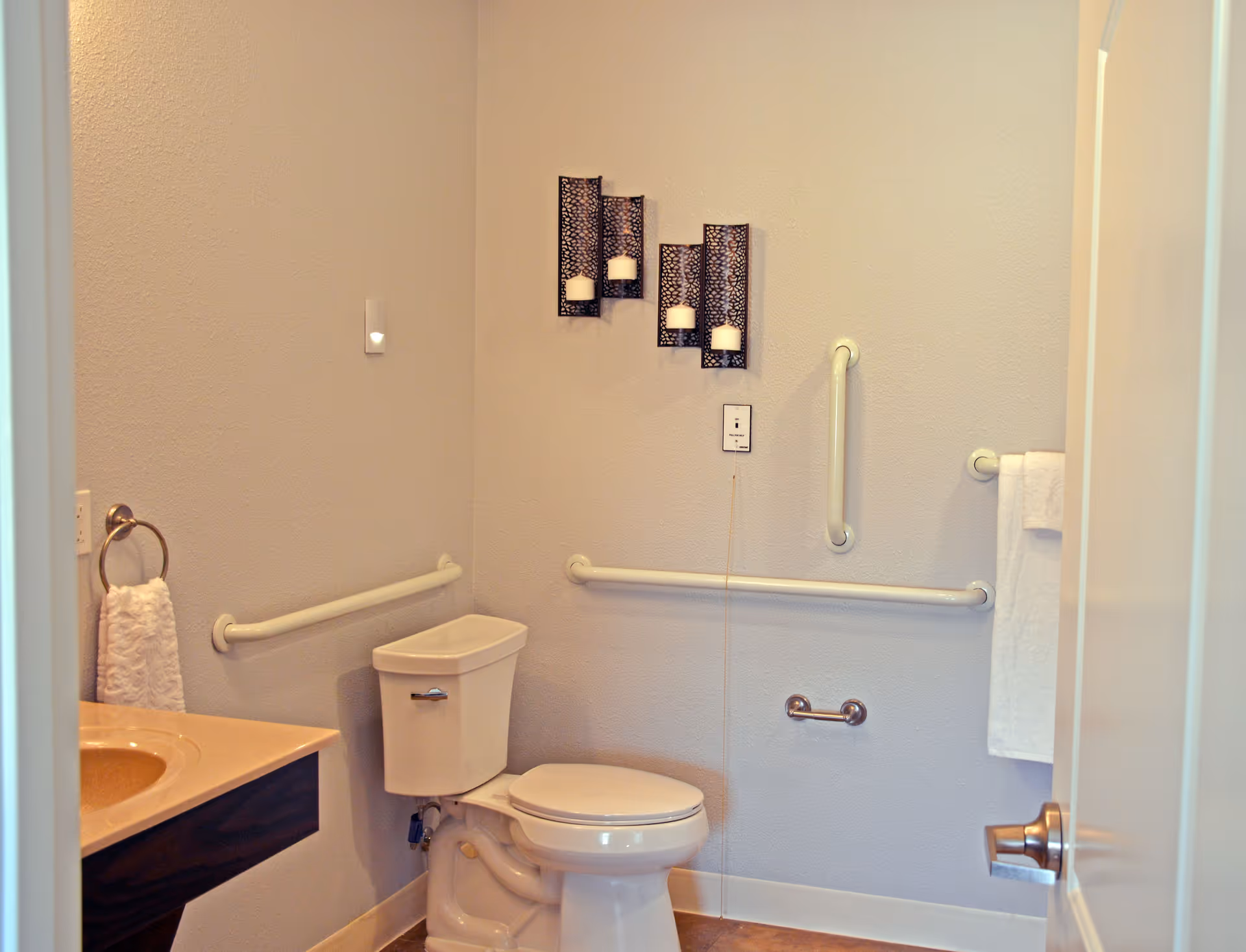 A clean and accessible bathroom with a toilet, grab bars on the walls, a towel ring with a white towel, and decorative candle holders mounted on the wall.