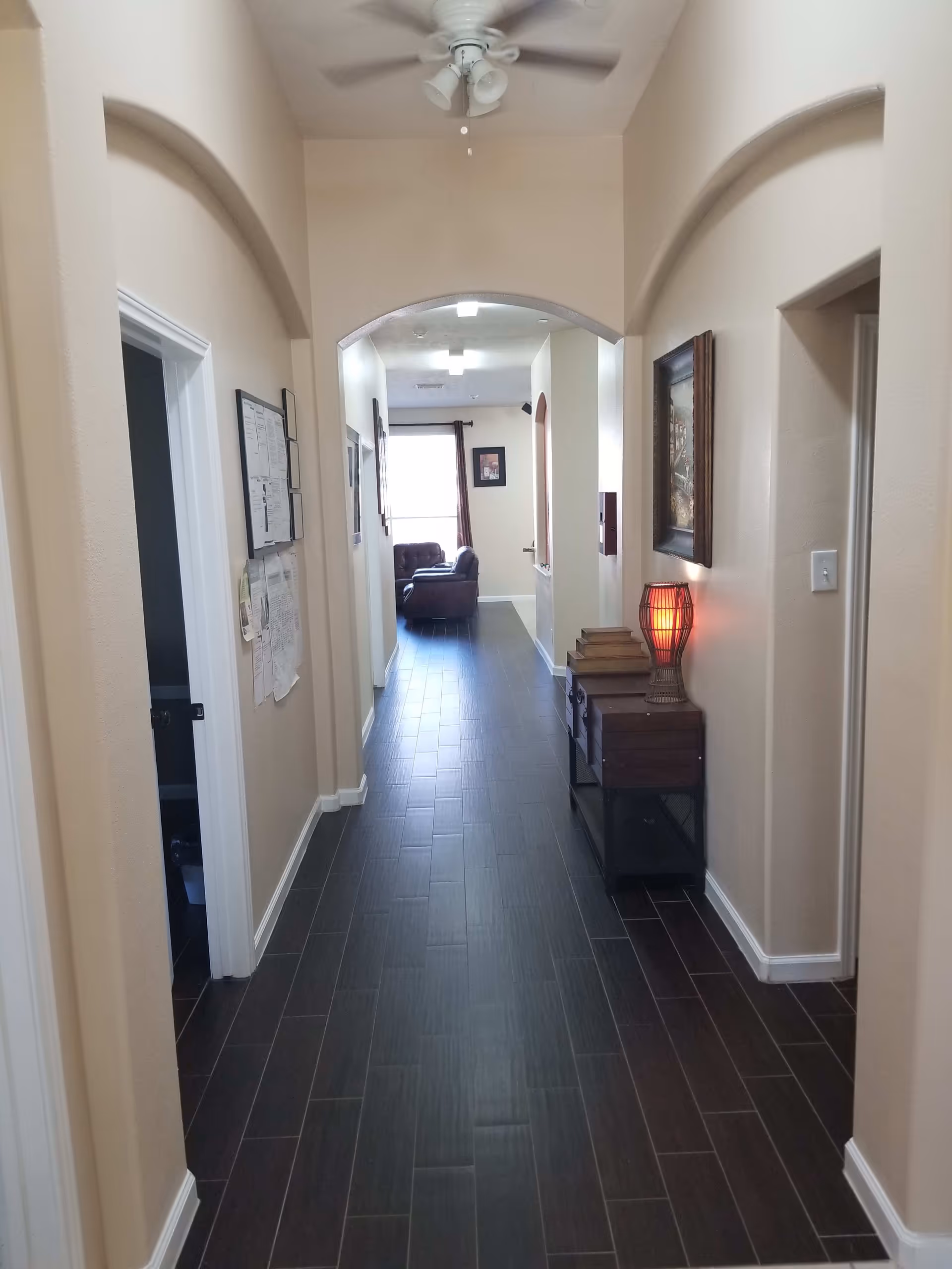 A long hallway with dark wood flooring and beige walls. On the right side, there is a small table with stacked books and a red lamp. A framed picture hangs above the table. The hallway leads to a room with a window and a brown armchair visible in the distance. The ceiling has a white fan with lights.