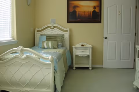 A small bedroom with a single white bed, matching nightstand, framed artwork, and a closed door marked '3'.