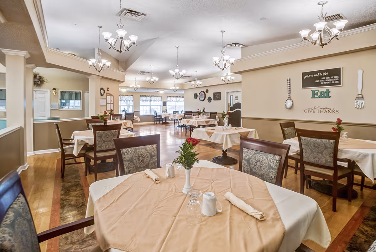 A spacious dining room with multiple tables covered in beige tablecloths and set with napkins, glasses, and small flower vases. The room features wooden chairs with patterned cushions, several chandeliers hanging from the ceiling, and large windows letting in natural light. Wall decor includes a sign with the word 'Eat' and utensils mounted on the wall.