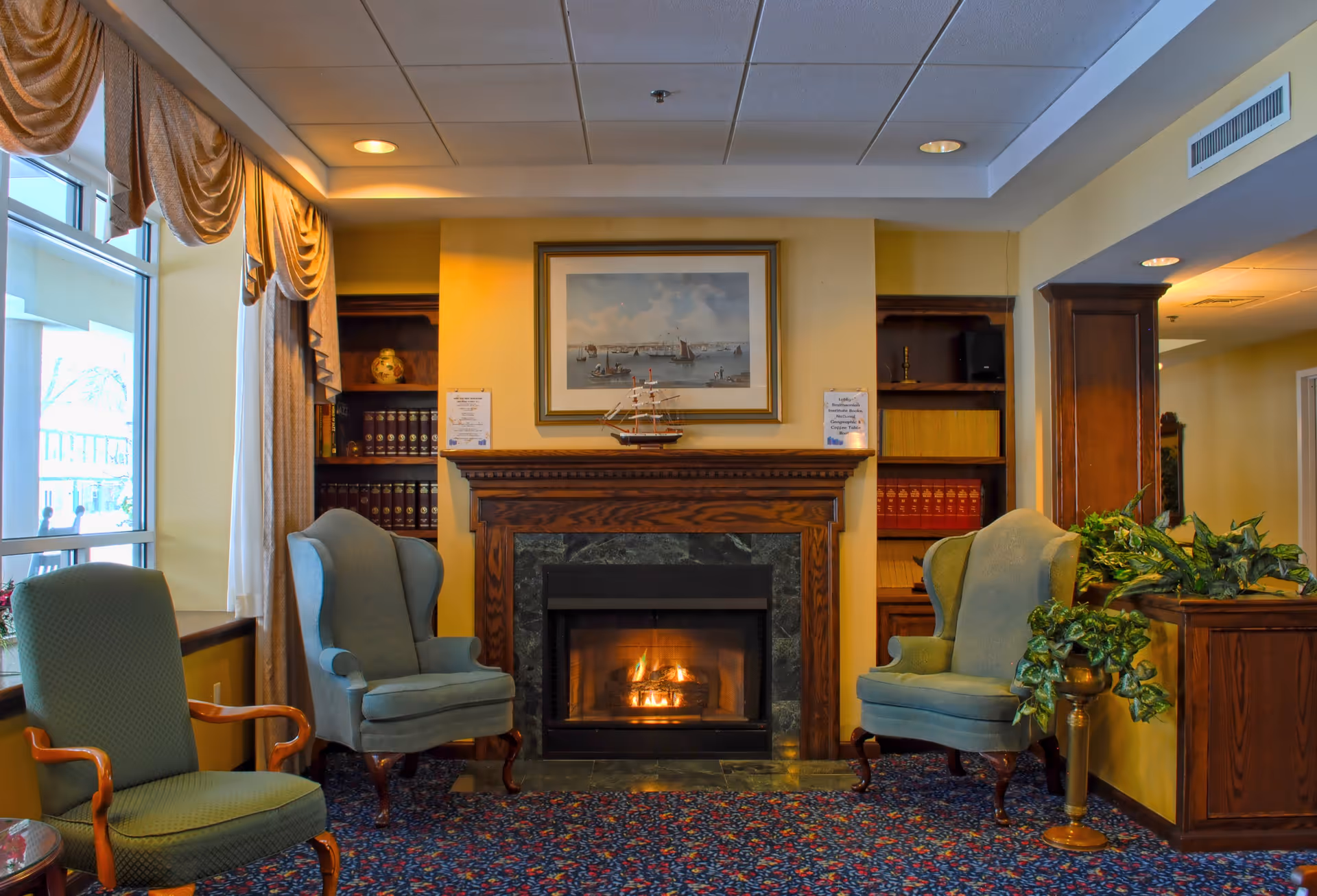 A cozy living room area with a lit fireplace surrounded by a wooden mantel. Two blue upholstered armchairs are positioned on either side of the fireplace, and a green armchair is placed near a window with draped curtains. Built-in wooden bookshelves flank the fireplace, holding books and decorative items. A framed painting of sailboats hangs above the mantel, and a model ship is displayed on it. There is a patterned carpet on the floor and some green plants near the right armchair.