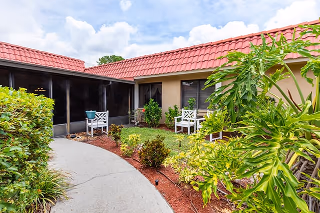 Outdoor courtyard area at Brookdale Bradenton Gardens with a curved concrete pathway, green shrubs, and plants. There are white chairs placed near the building with a red tiled roof and beige walls under a partly cloudy sky.