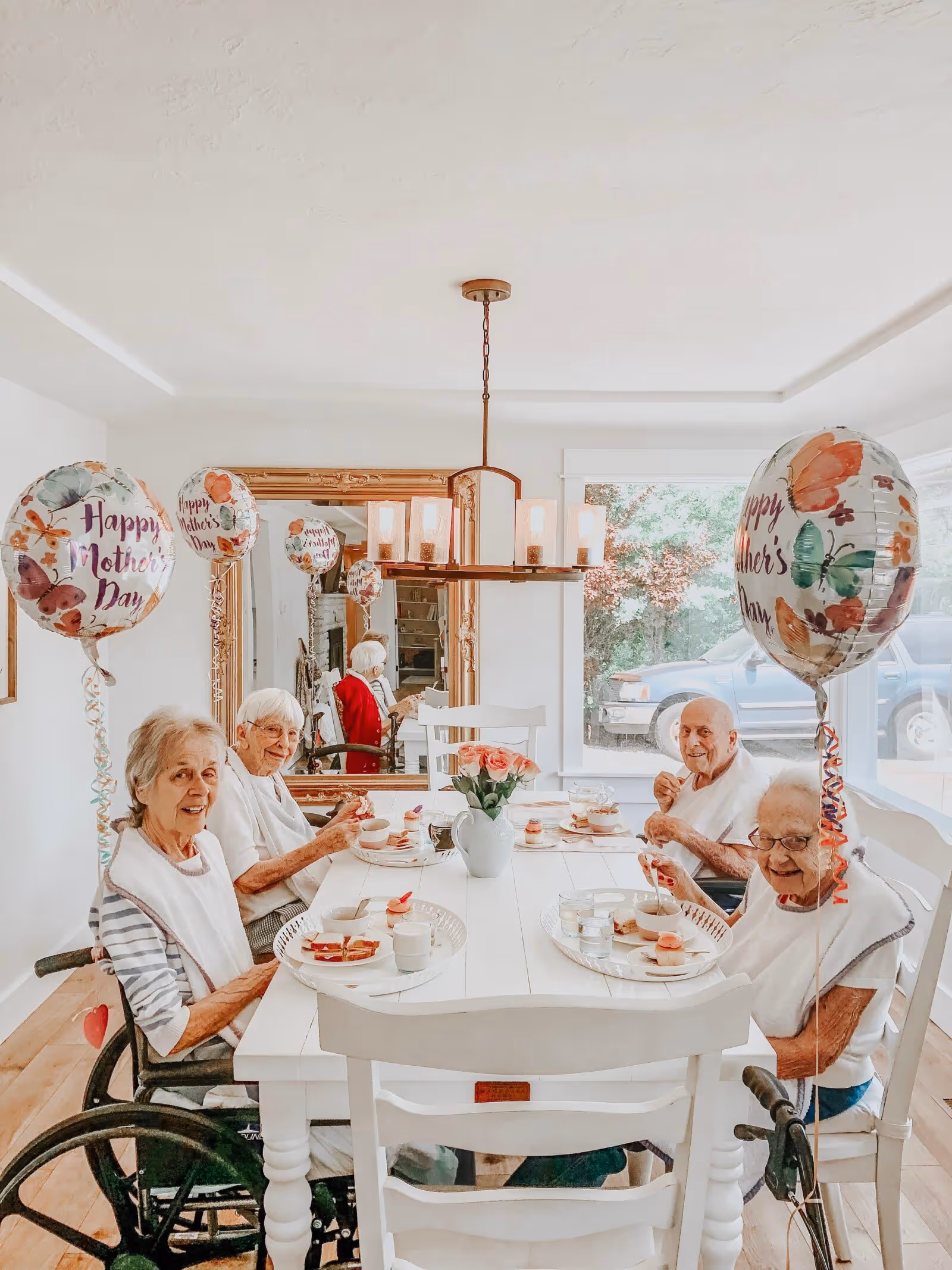 Four elderly people sitting around a white dining table in a bright room, enjoying a meal together. Each person has a plate with food and a glass of water. There are three balloons with 'Happy Mother's Day' written on them, and a vase with pink roses is placed in the center of the table. A large mirror and a window showing a parked vehicle are visible in the background.