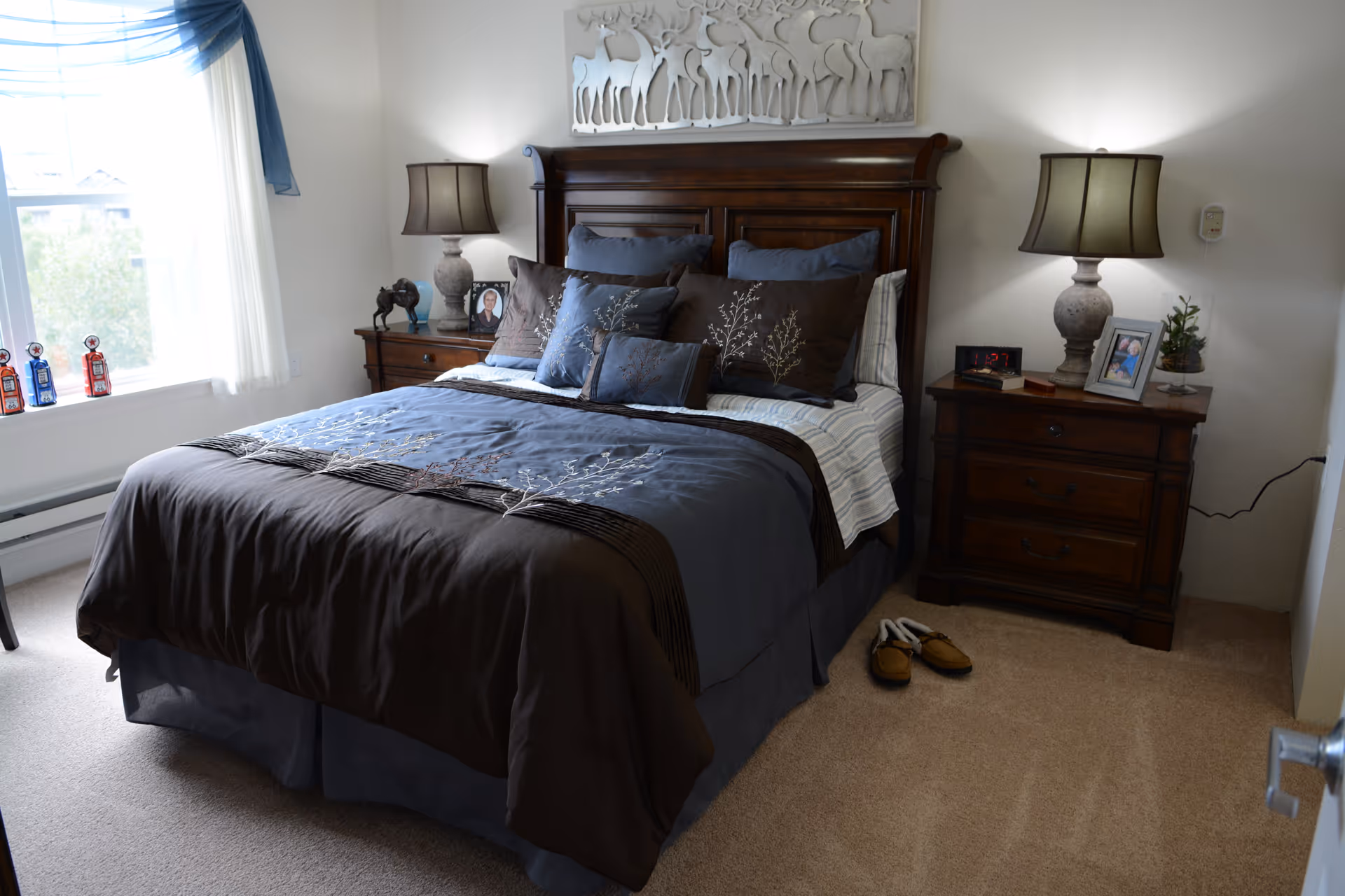 A neatly made bed with blue and brown bedding in a bedroom featuring a wooden headboard. There are two matching wooden nightstands on either side of the bed, each with a lamp. One nightstand has a framed photo and a small plant, while the other has a framed photo and a decorative horse figurine. A pair of slippers is placed on the carpeted floor next to the bed. A window with white curtains and a blue valance allows natural light into the room. Above the bed is a decorative wall art piece with a deer motif.