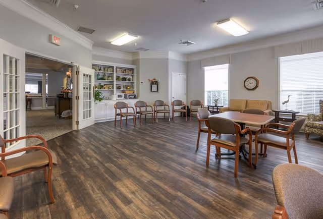 A spacious common area in a senior living facility with wood flooring, several chairs arranged along the walls and around tables, large windows with blinds, a wall clock, built-in shelves with decorative items, and double glass doors leading to another room.
