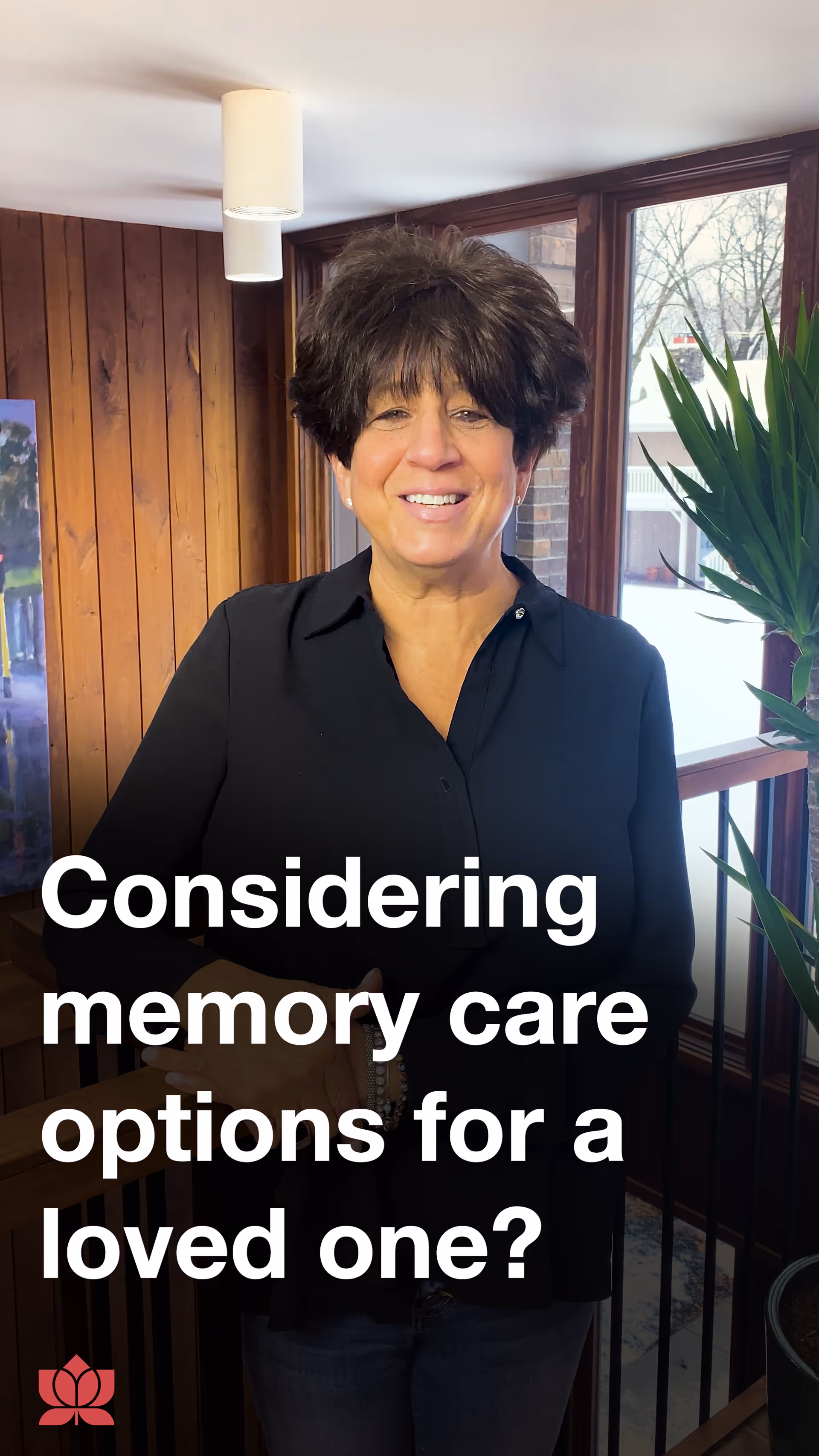 A smiling woman with short dark hair wearing a black shirt stands indoors in front of a window with wooden paneling on the wall behind her. There is a green plant to the right and a white ceiling light above. Text on the image reads 'Considering memory care options for a loved one?'