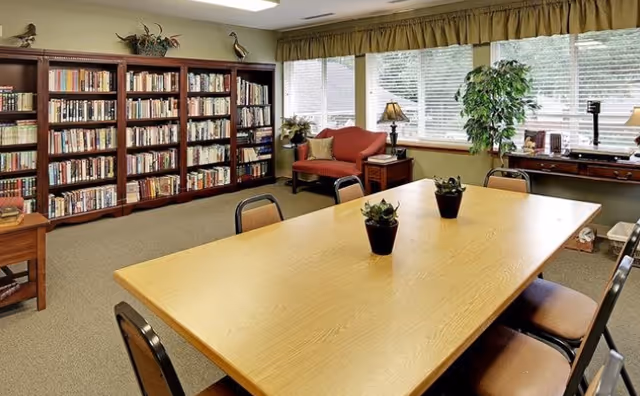 A cozy senior living common room with a large wooden table surrounded by chairs, three small potted plants on the table, a bookshelf filled with books along one wall, a red loveseat with a pillow, a side table with a lamp, a large window with blinds and curtains, and a desk with a plant and other items.