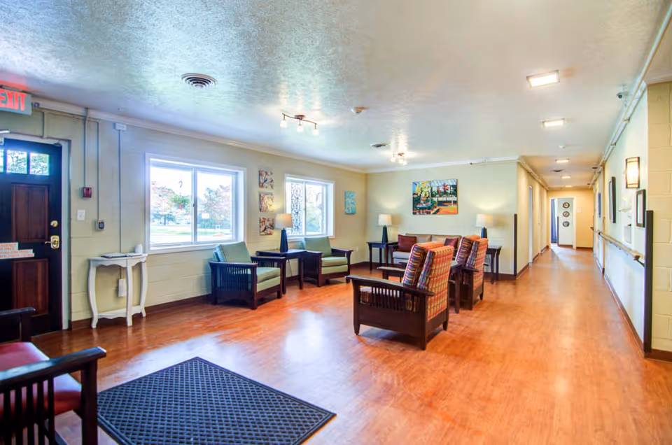 A bright and spacious senior living facility common area with wooden flooring, several armchairs arranged around small tables, two large windows letting in natural light, wall art, and lamps. A hallway extends to the right, and a dark wooden door with an exit sign is on the left.