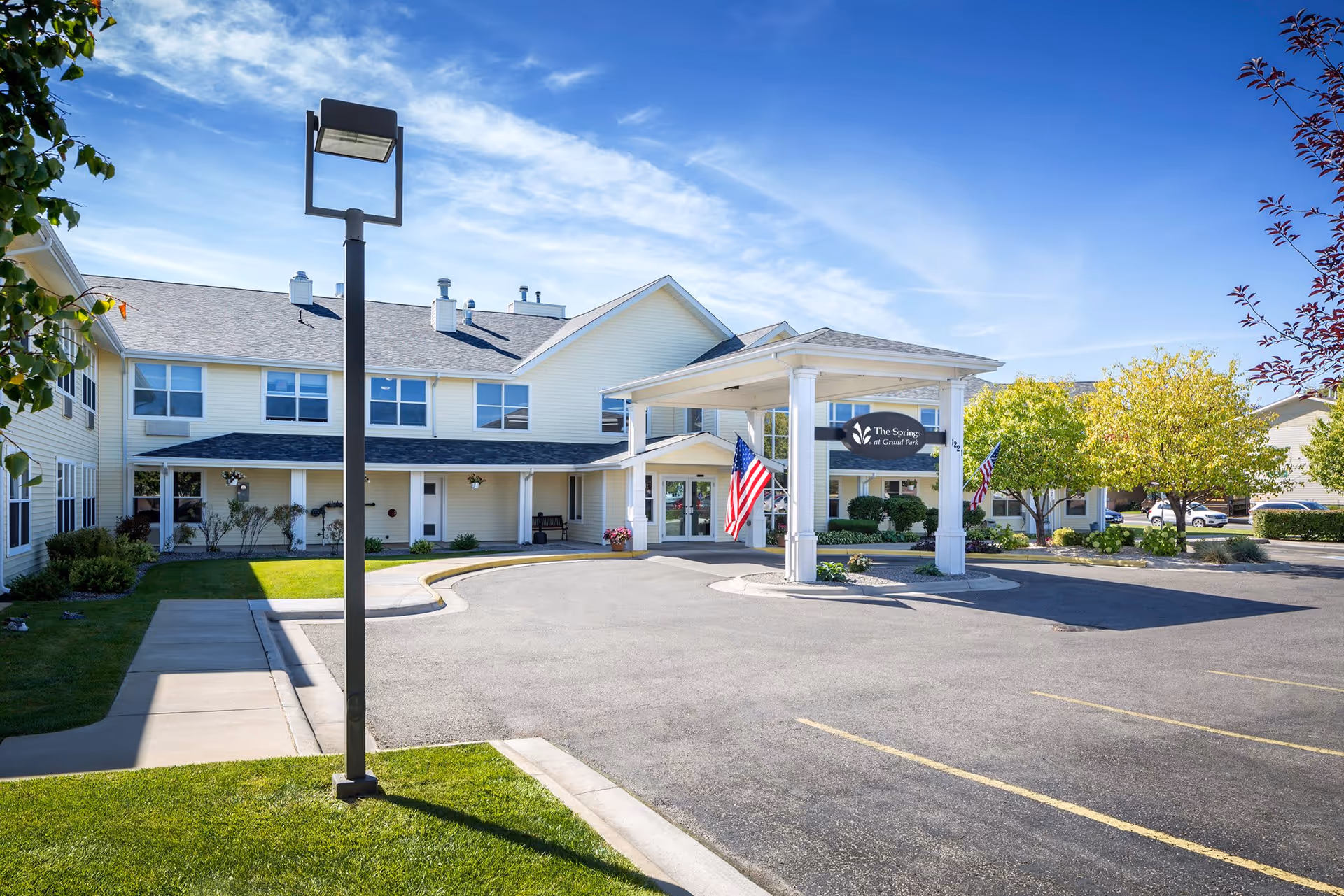 Exterior view of The Springs at Grand Park senior living facility showing a two-story building with a covered entrance, American flags, a parking lot, and landscaped greenery under a blue sky.