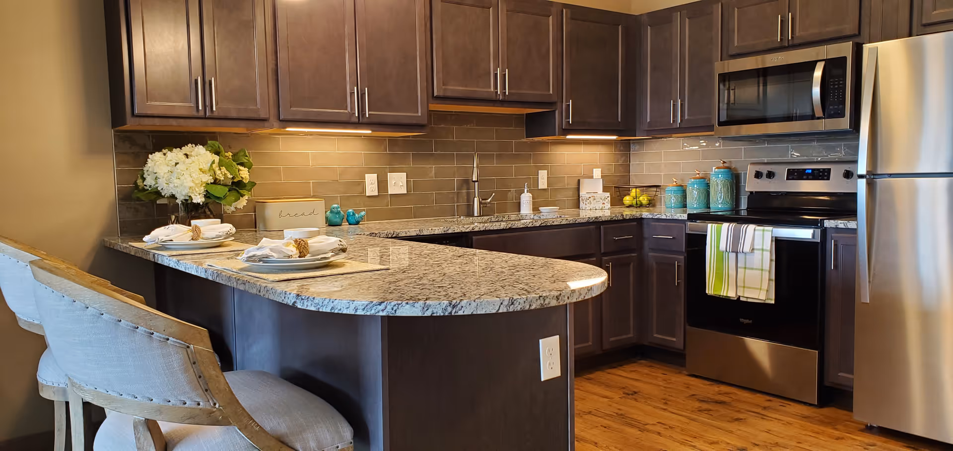 Modern kitchen with dark wooden cabinets, granite countertops, stainless steel appliances including a refrigerator, stove, and microwave. The kitchen island has two cushioned bar stools and is set with plates and napkins. There are decorative items such as a vase with white flowers, a bread box, and ceramic jars on the countertop.