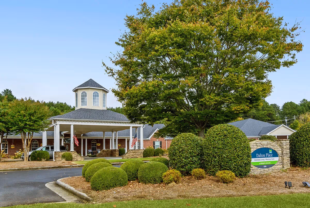 Exterior view of Dalton Place assisted living and memory care facility showing a brick building with a covered entrance, manicured bushes, a large tree, and a sign with the facility name.
