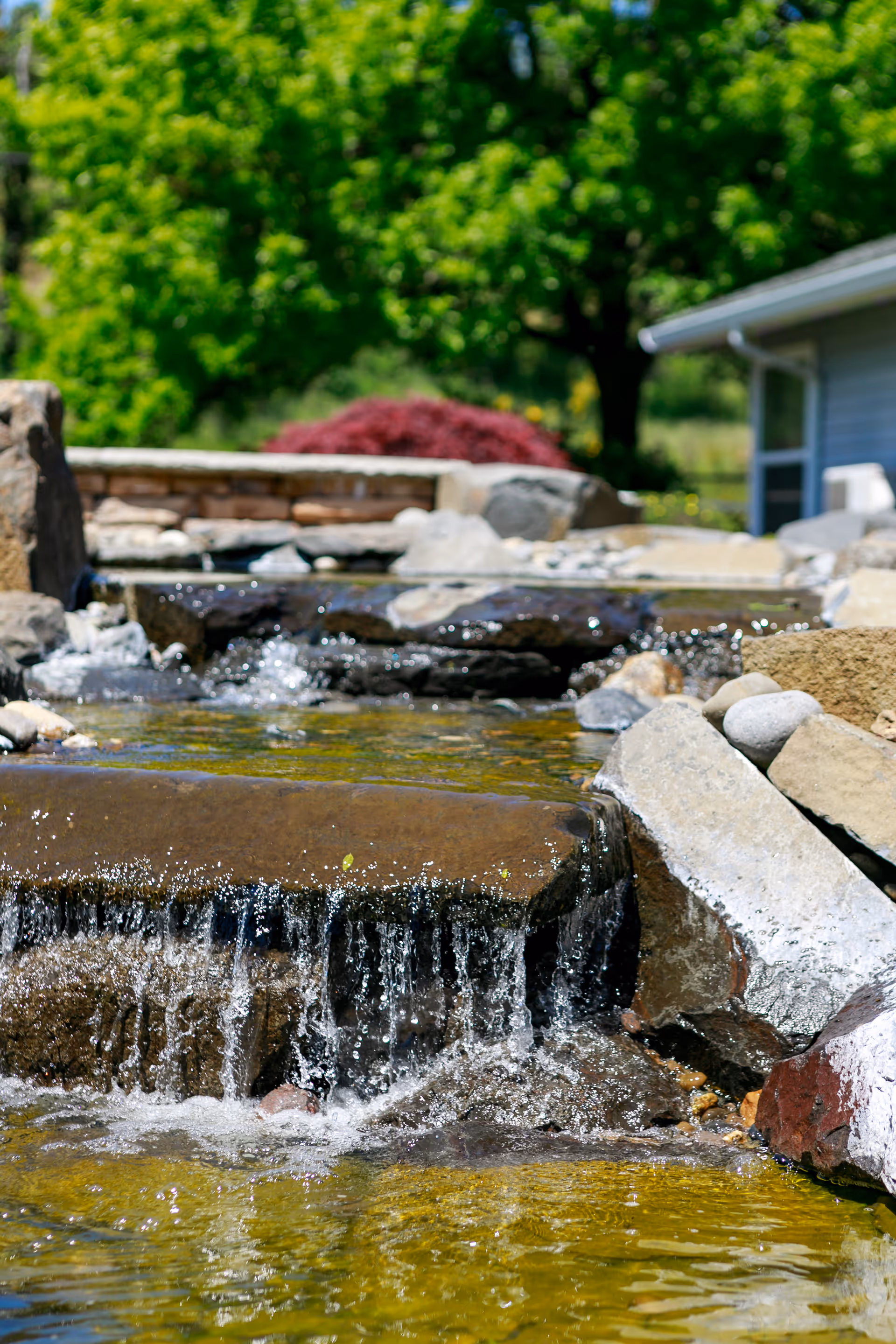 A small cascading water feature with clear water flowing over flat rocks surrounded by larger stones, with green trees and a building partially visible in the background.