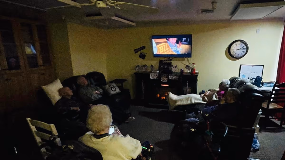 A dimly lit living room with several elderly people seated in recliners and chairs, watching a television mounted on the wall above a fireplace. The room has a ceiling fan, a clock on the wall, and a cabinet with glass doors on the left side.