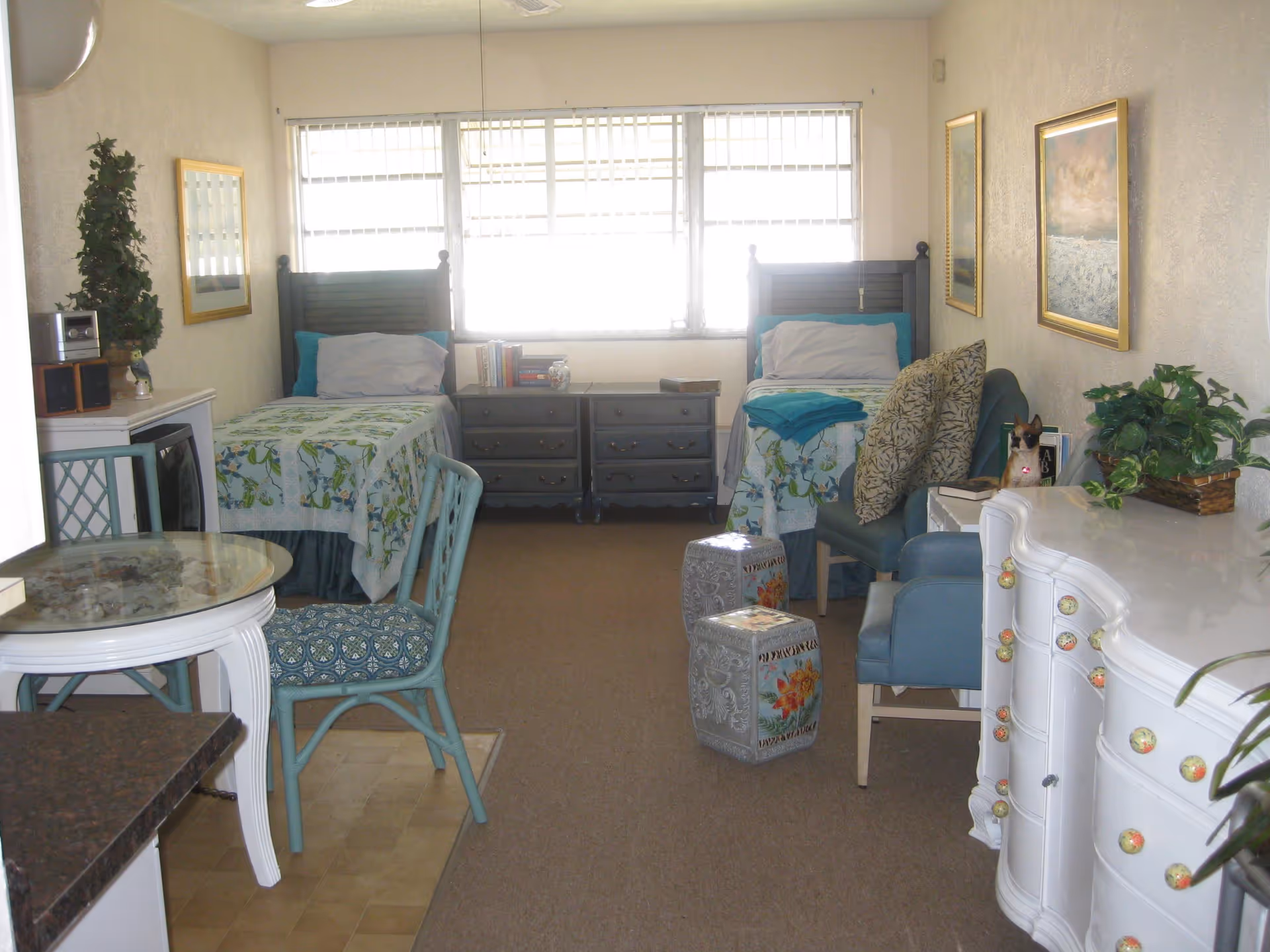 A bright room with two single beds positioned side by side against a window with blinds. Each bed has a floral bedspread and pillows. Between the beds are two dark wooden nightstands with books and a decorative item. In the foreground, there is a small round glass-top table with two blue chairs. On the right side, there is a white dresser with colorful knobs, a blue armchair with patterned cushions, and decorative plants and artwork on the walls.