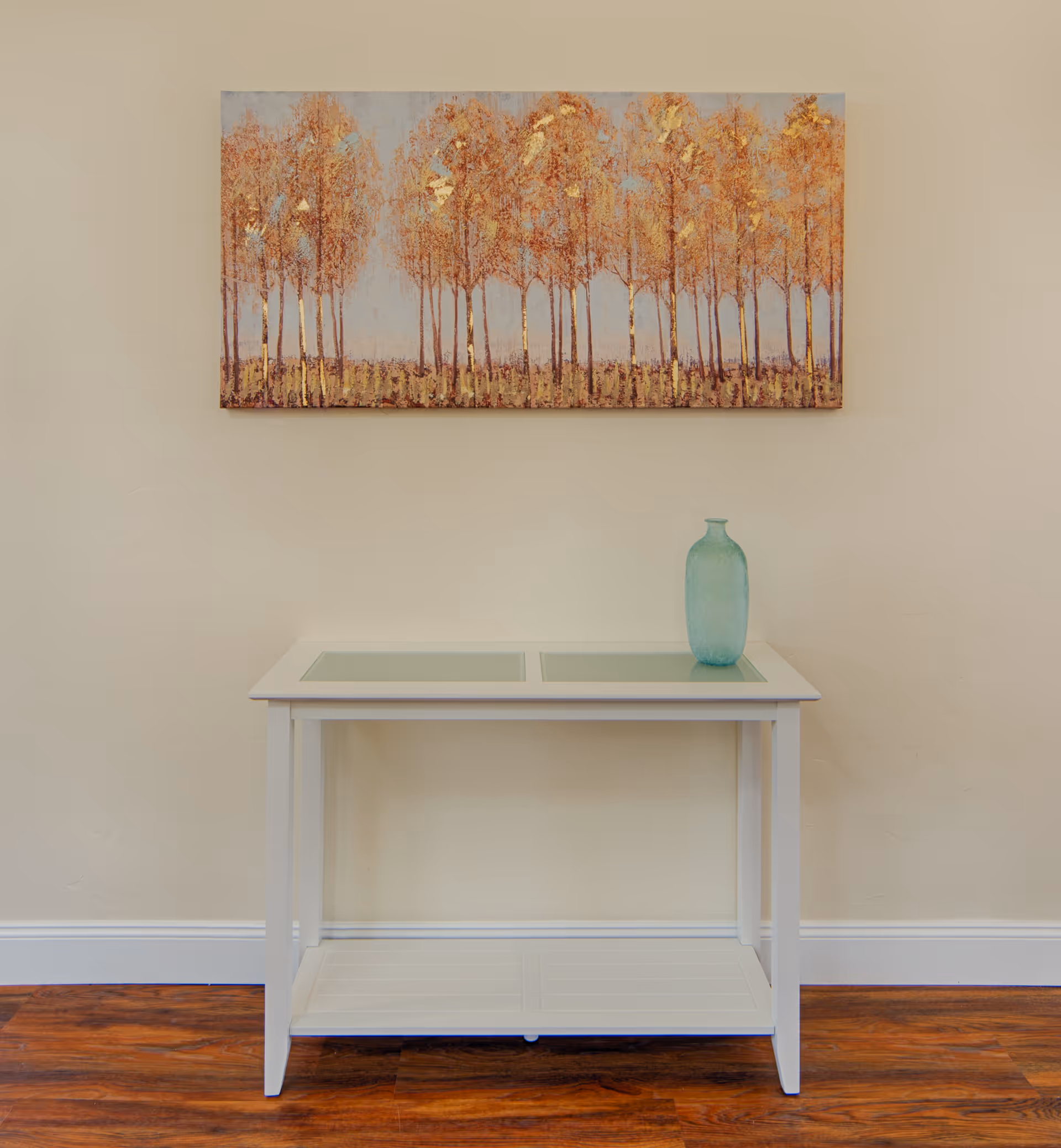 A white console table with glass panels on top and a lower shelf, placed against a beige wall with a wooden floor. On the table is a single blue-green decorative vase. Above the table hangs a rectangular painting depicting a row of tall trees with golden and orange leaves against a light blue sky.