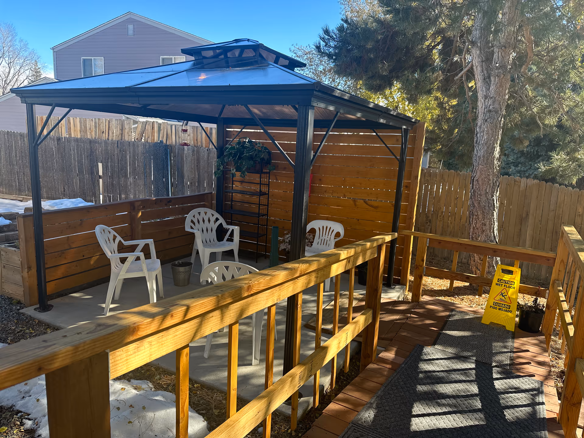 Outdoor seating area with a metal gazebo covering four white plastic chairs arranged on a concrete patio. The area is enclosed by wooden fencing and has a wooden railing along a ramp leading to the seating area. There is a yellow caution wet floor sign on the ramp, and trees and a house are visible in the background.