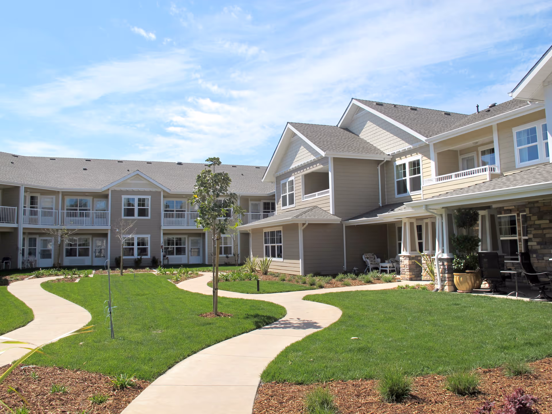 Outdoor view of Camden Springs Gracious Retirement Living facility showing a well-maintained courtyard with green grass, young trees, curved concrete walkways, and a two-story beige building with white trim and balconies under a blue sky with some clouds.