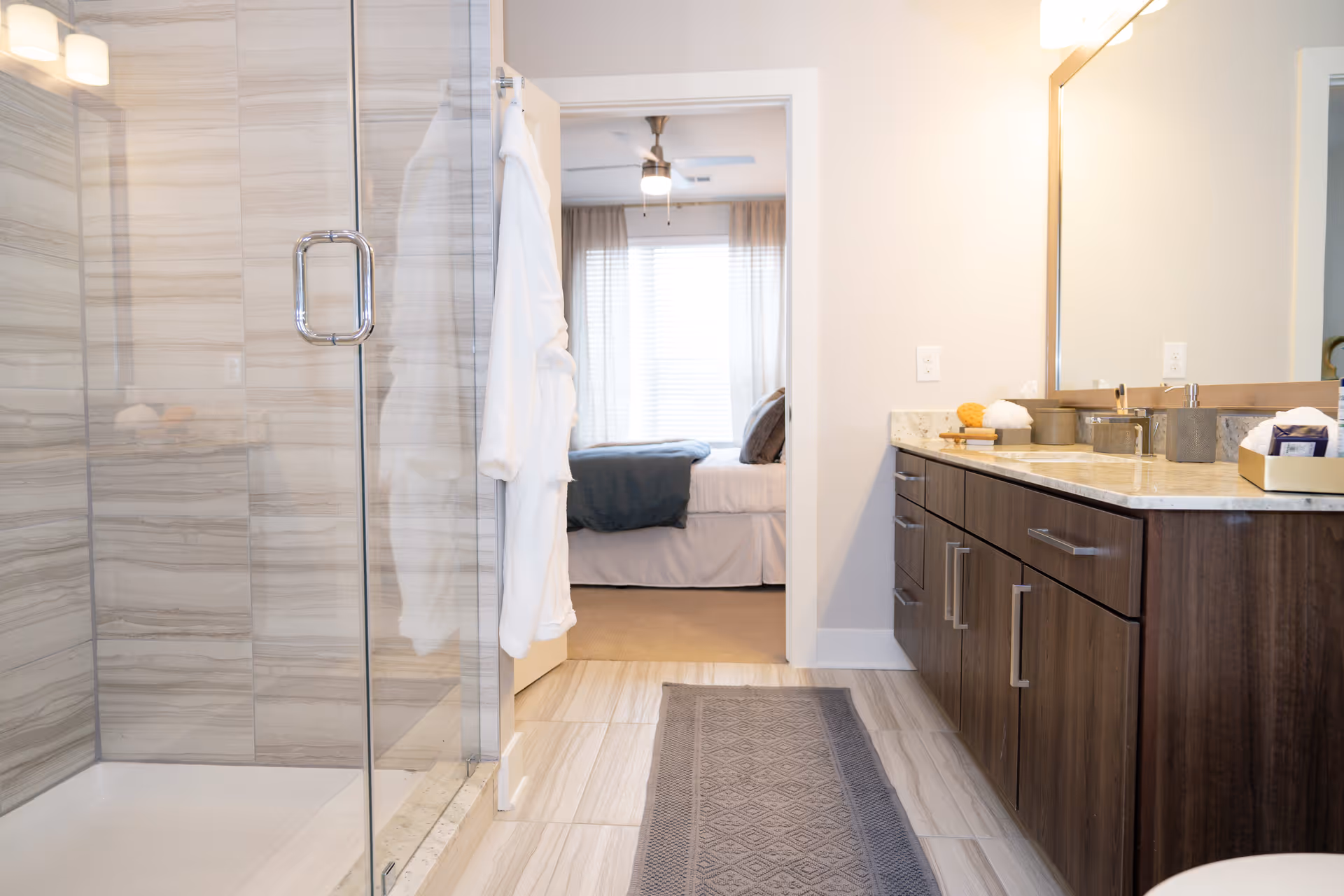 A modern bathroom with a glass-enclosed shower on the left, a white bathrobe hanging on the door, a long wooden vanity with a marble countertop and a large mirror on the right. Through the open door, a bedroom with a bed, pillows, and a ceiling fan is visible.