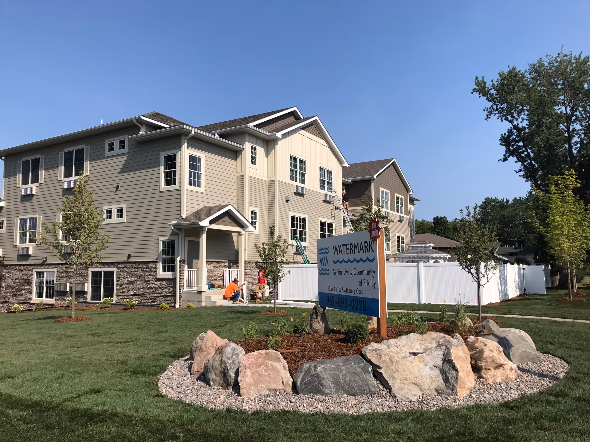 Exterior view of a multi-story senior living facility building with beige and brown siding, multiple windows, and a small porch entrance. In front of the building is a landscaped area with rocks, small trees, and a sign that reads 'WATERMARK Senior Living Community of Fridley Care Suites & Memory Care 952-893-0228'. The sky is clear and blue.