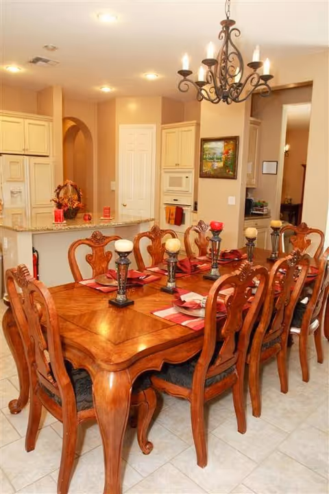 Formal dining room with an ornate wooden table set for eight, a chandelier overhead, and a view into a kitchen area.