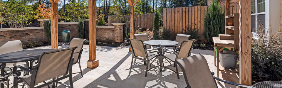 Outdoor patio area with multiple round tables and chairs under a wooden pergola, surrounded by a wooden fence and landscaped plants, with sunlight casting shadows on the concrete floor.