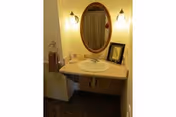 A bathroom sink area with a beige countertop, an oval mirror mounted on the wall, two wall-mounted light fixtures on either side of the mirror, a towel ring with a brown towel, and a small framed picture placed on the countertop.