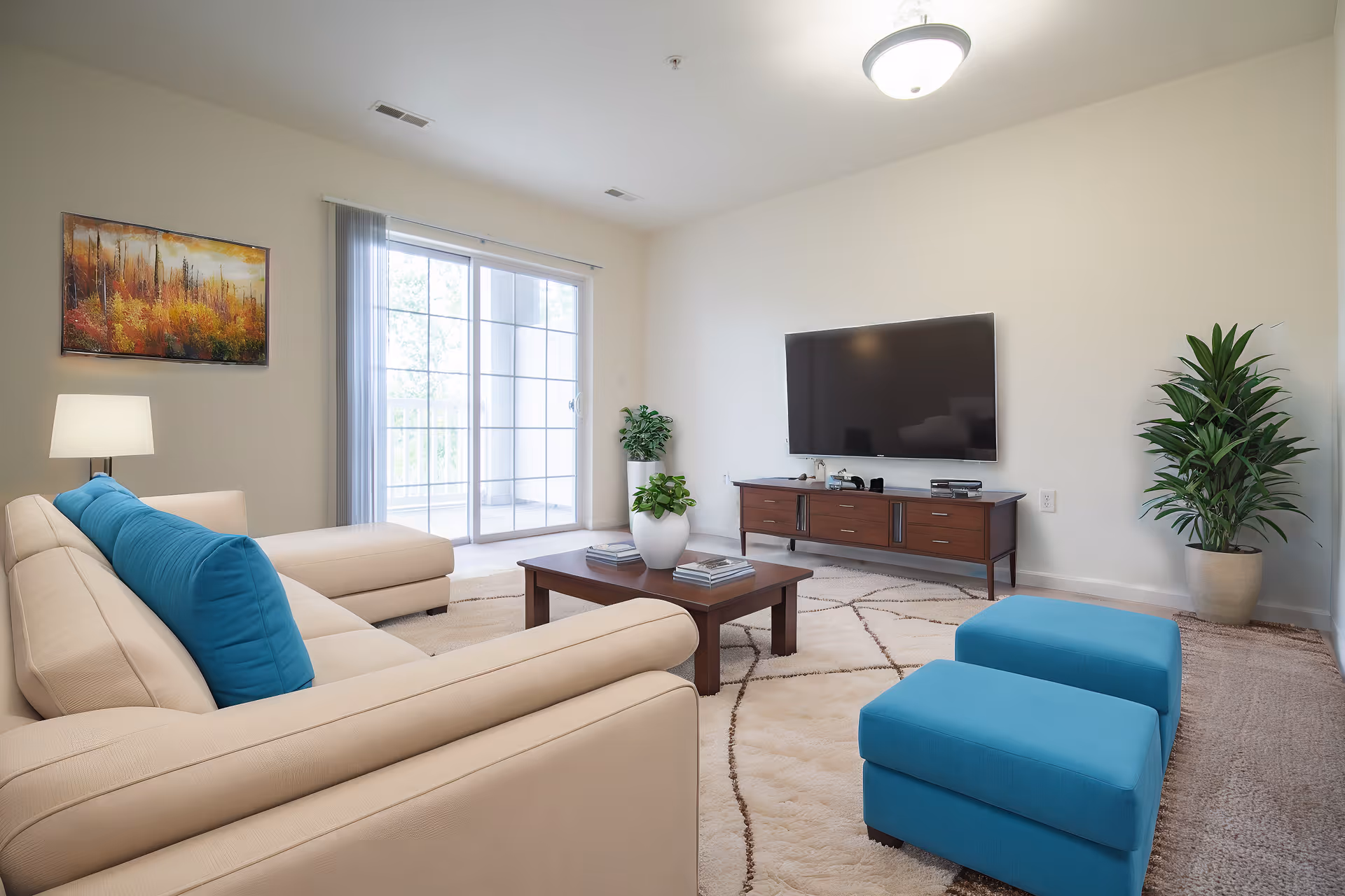 Bright living room with a beige sectional sofa, blue ottomans and pillows, a coffee table, wall-mounted TV, and sliding glass doors to a balcony.