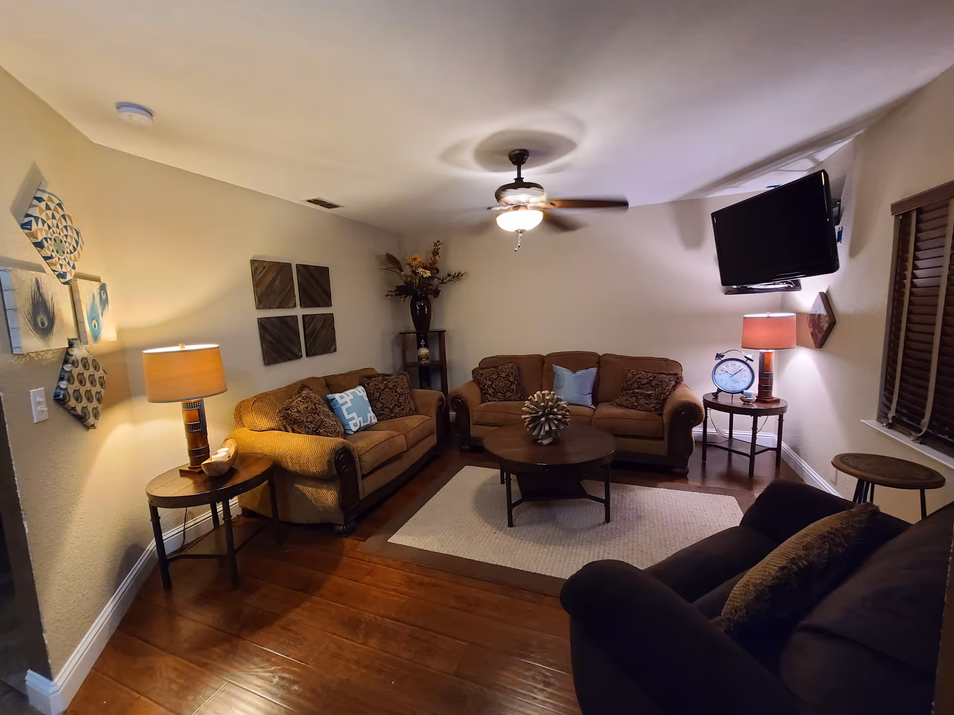 A cozy living room with two brown sofas and one dark armchair arranged around a round wooden coffee table on a beige rug. The room has wooden flooring, a ceiling fan with a light, two side tables with lamps, wall decorations, a large vase with flowers in the corner, and a wall-mounted flat-screen TV.