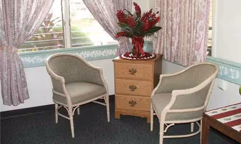 Two wicker chairs flank a small dresser with a red plant in a windowed, curtained lounge.
