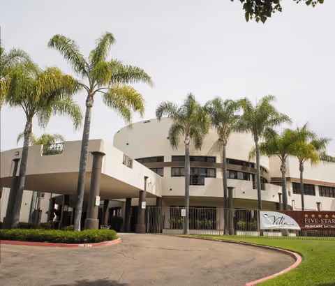 Exterior view of Villa Rancho Bernardo Care Center, a two-story building with a curved facade, surrounded by palm trees and a driveway with a covered entrance.