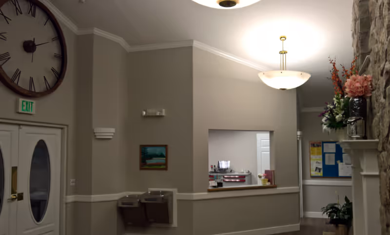Interior view of a senior living facility lobby or common area with beige walls, a large wall clock above double doors, a water fountain, a small painting, a reception window, a bulletin board, and floral arrangements on a stone fireplace mantel.