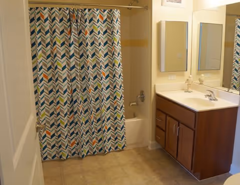 Bathroom with a patterned shower curtain over a tub, a vanity with sink and mirror, and a tiled floor.
