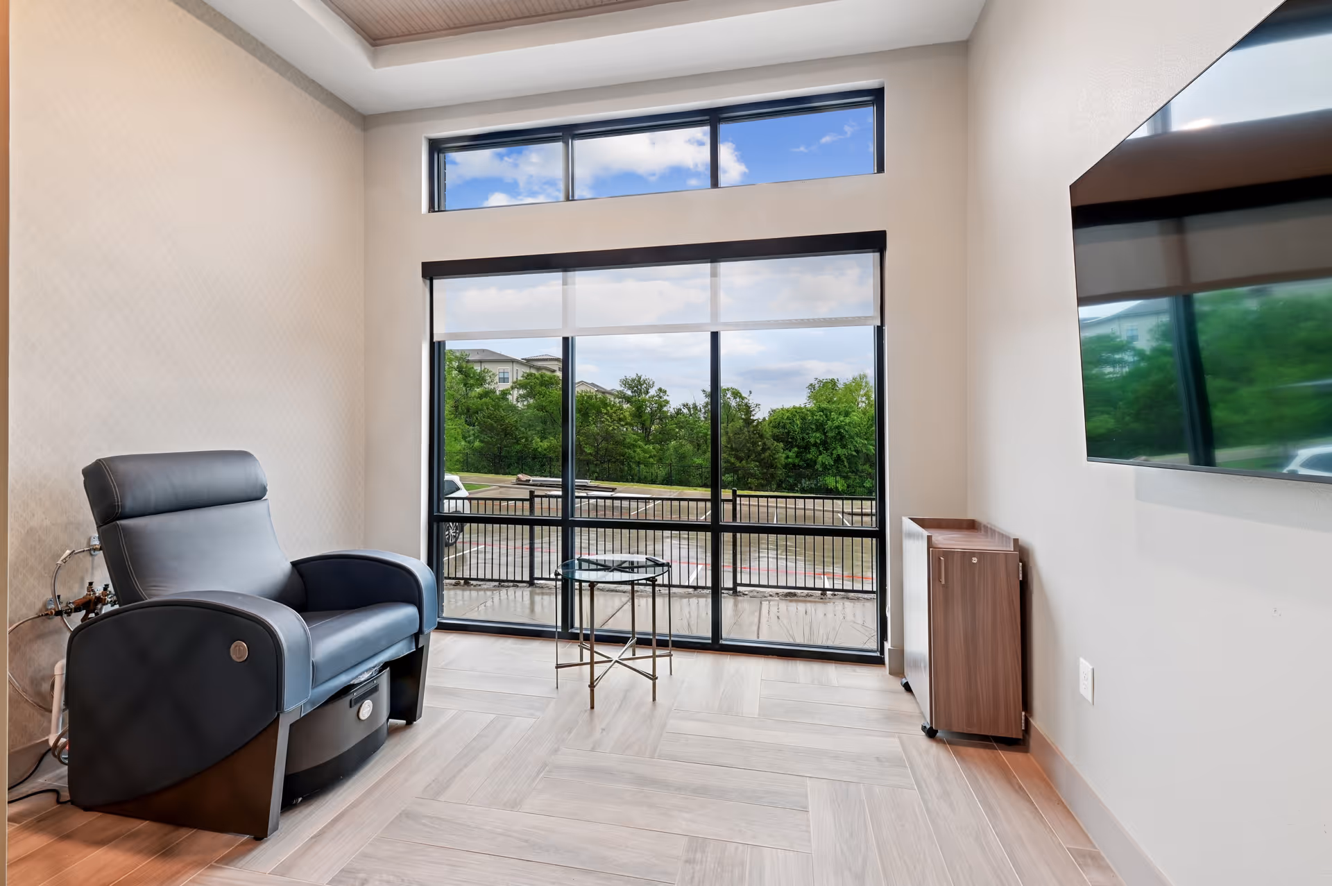 A modern room with a large window and glass door overlooking a parking lot and greenery outside. Inside, there is a black leather recliner chair, a small round glass side table, a wall-mounted flat screen TV, and a wooden cabinet on wheels. The floor has light wood-colored tiles arranged in a herringbone pattern.