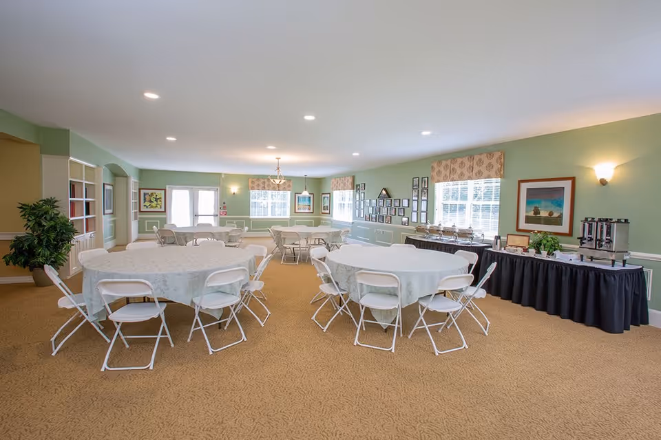 A spacious dining room with round tables covered in light-colored tablecloths and surrounded by white folding chairs. The room has green walls with white trim, several windows with patterned valances, framed artwork on the walls, and a buffet table with coffee urns and serving dishes along one side. The carpet is beige, and the ceiling has recessed lighting.