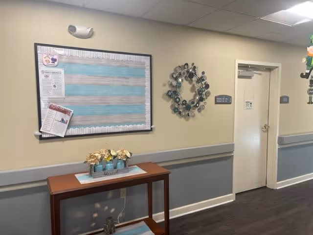 A hallway in a senior living facility with beige and gray walls, a wooden table holding three small flower pots, a decorative metal wall wreath, a bulletin board with papers pinned on it, and a closed white door with a sign next to it.