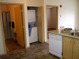 Interior view of a senior living facility kitchen area with a dishwasher, sink, and countertop on the right. In the background, there is a laundry area with a stacked washer and dryer, and an open door leading to a bathroom with a shower curtain visible.