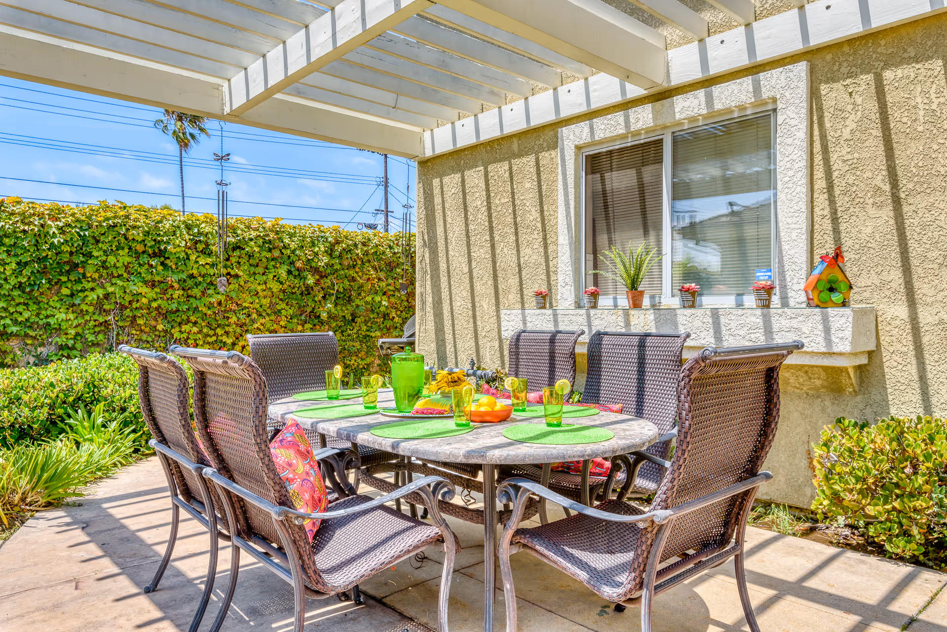 Outdoor covered patio with a round dining table set for six, wicker chairs, potted plants, and a hedge in the background.