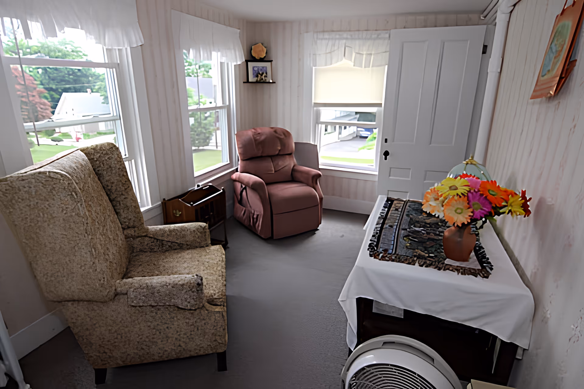 Cozy sitting room with two upholstered armchairs by windows, a small table with a vase of colorful flowers, and an entry door.