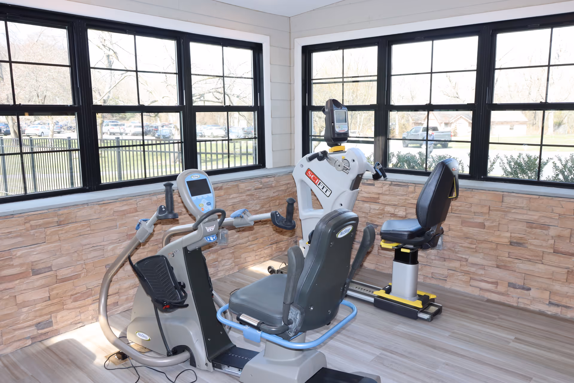 A bright exercise room with large windows showing an outdoor view. The room contains two pieces of exercise equipment designed for seated use, including a recumbent bike and an arm and leg exercise machine. The walls have a stone-like texture and the floor is light wood.