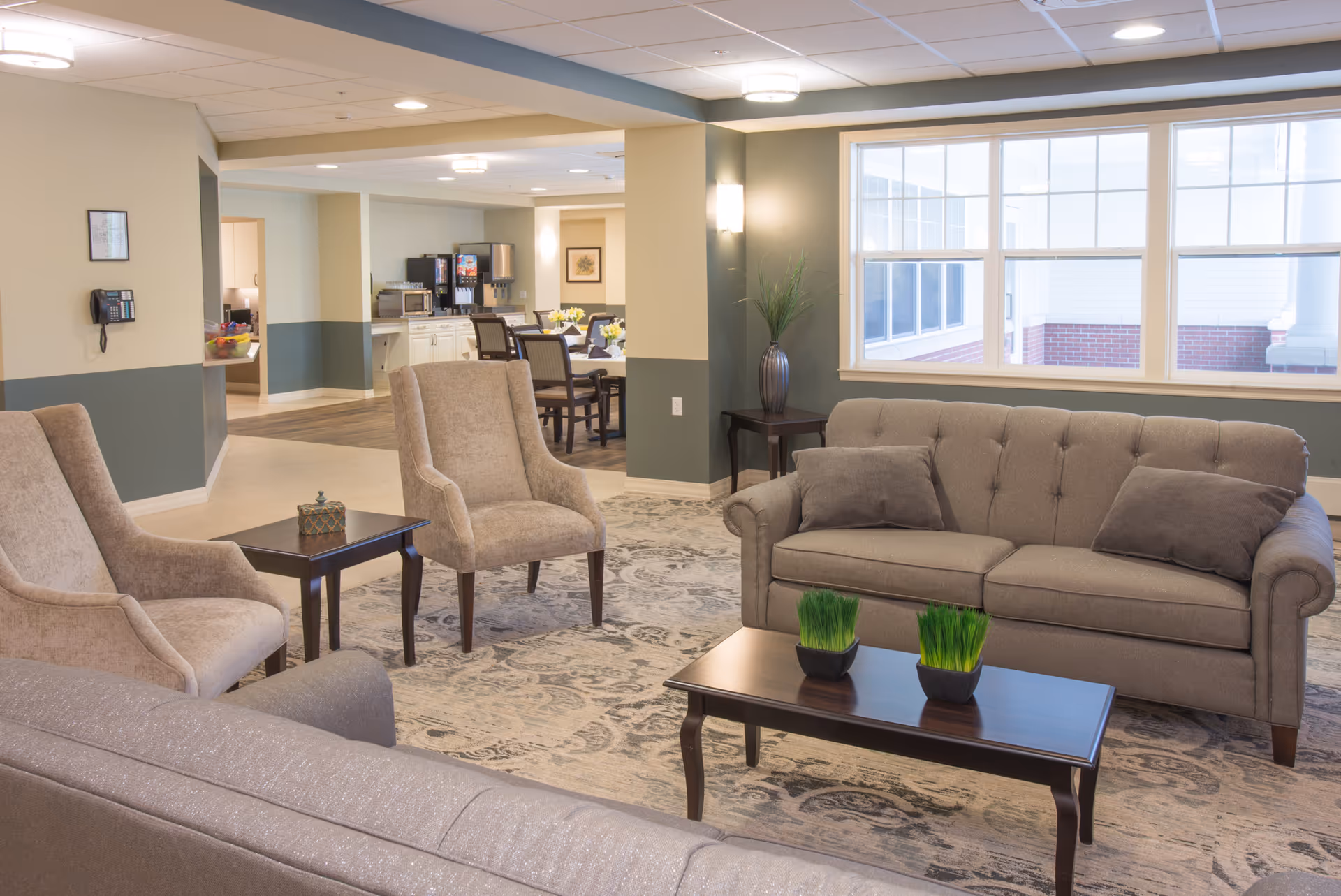 Bright senior living lounge with sofas and armchairs around a coffee table and a dining/service area visible in the background.