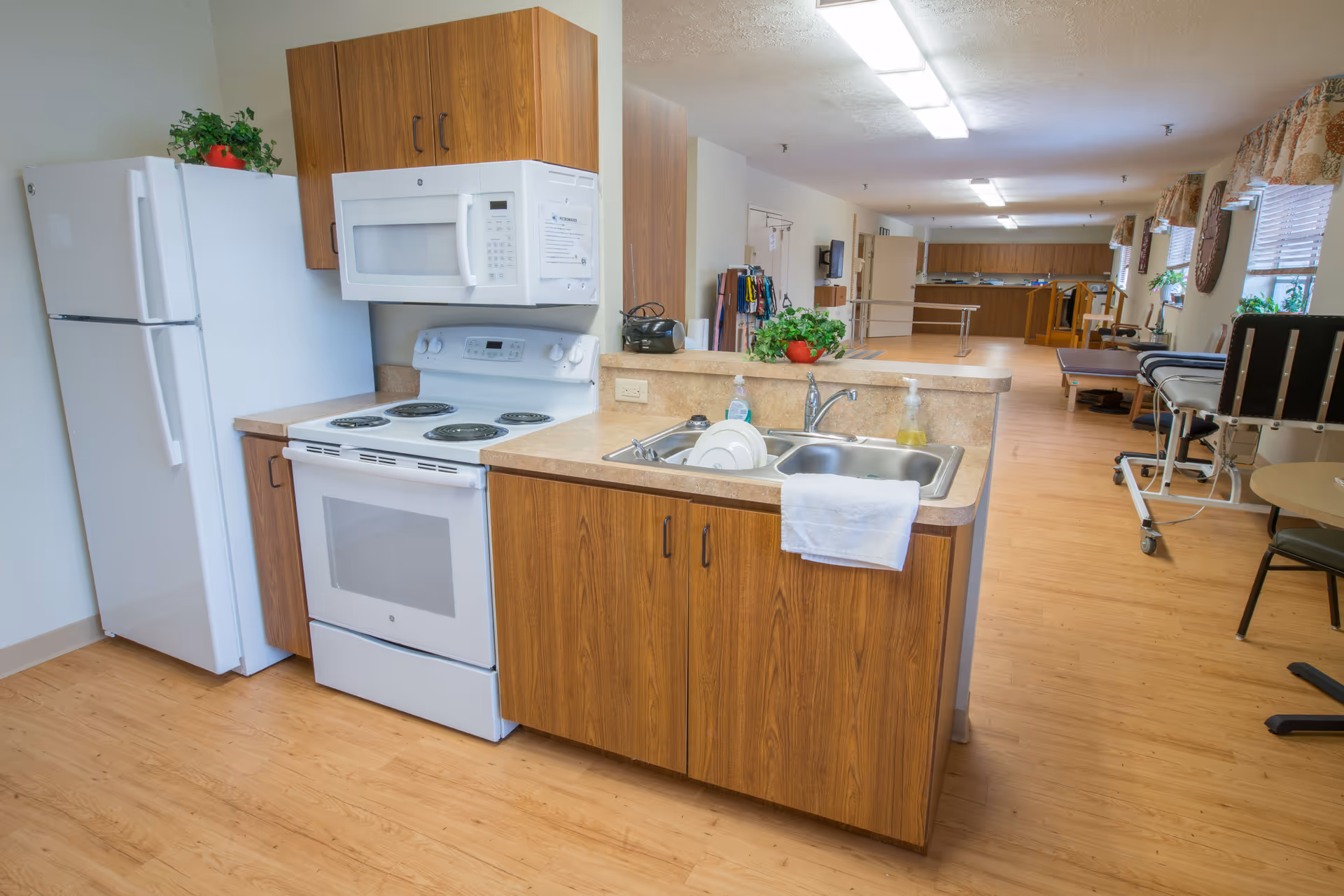 A kitchen area in a senior living facility with wooden cabinets, a white refrigerator, stove, and microwave. The kitchen counter has a double sink with dishes and a towel hanging on the edge. In the background, there is a spacious room with wooden flooring, windows with curtains, plants, and some medical or mobility equipment.